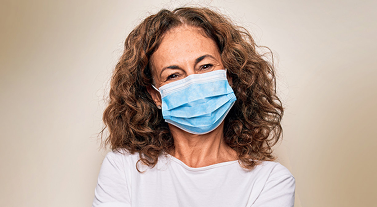 Caucasian woman with curly brown hair and a mask on her face, against a tan background